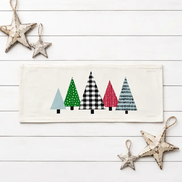 A rectangular cream fabric runner or placemat featuring a row of stylized Christmas trees in different patterns (solid light blue, green with white dots, black and white gingham, red with white stripes, and blue-gray with white stripes). The item is displayed on a white wood plank background with rustic distressed metal star ornaments.
