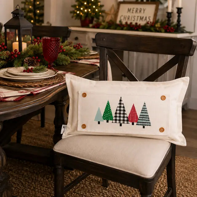 A rectangular cream throw pillow with a Christmas tree design, resting on a wooden dining chair in a decorated holiday dining room.