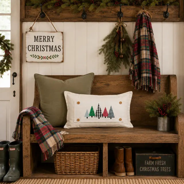 A decorative rectangular throw pillow with a Christmas tree pattern sits on a rustic wooden bench in a cozy holiday entryway. The bench is styled with other pillows, a plaid throw blanket, a hanging 'Merry Christmas' sign, greenery, and boots.