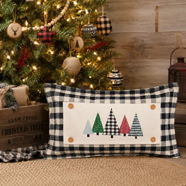 A rectangular holiday throw pillow with a black and white buffalo plaid border and a central white panel featuring four colorful Christmas tree silhouettes, styled in front of a decorated Christmas tree.