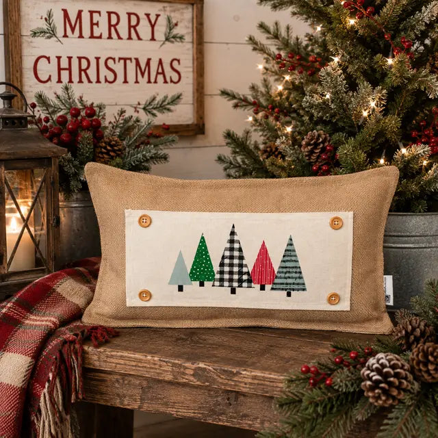 A rectangular burlap throw pillow with a cream fabric panel featuring four stylized Christmas trees in green, black-and-white plaid, red, and gray, displayed on a rustic wooden bench in a holiday setting with a lit Christmas tree, lantern, and 'Merry Christmas' sign.