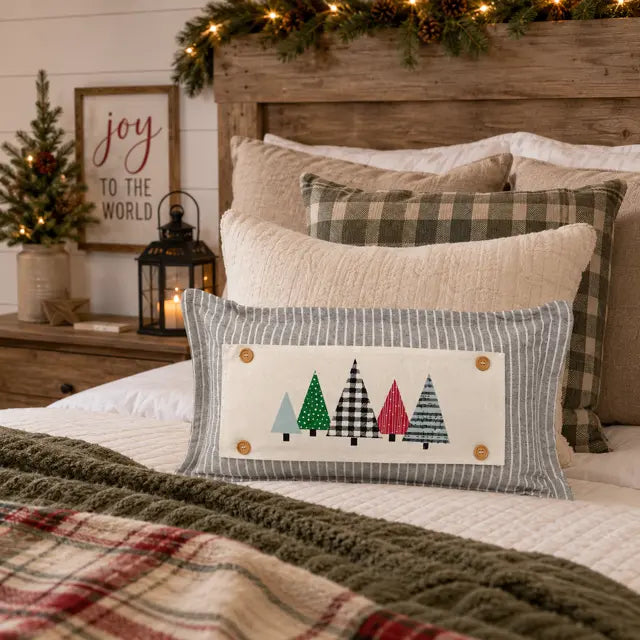 A rectangular decorative pillow with a striped gray border and a cream center panel featuring five colorful Christmas tree silhouettes, styled on a bed with other holiday pillows, a plaid blanket, and a wooden headboard in a cozy bedroom setting.
