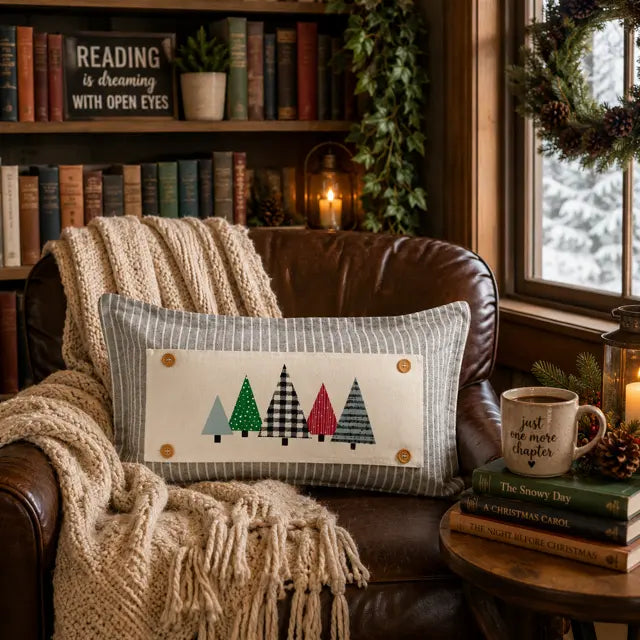 A rectangular throw pillow with a striped gray and white border and four colorful pine tree designs, resting on a brown leather armchair in a cozy, book-filled room.