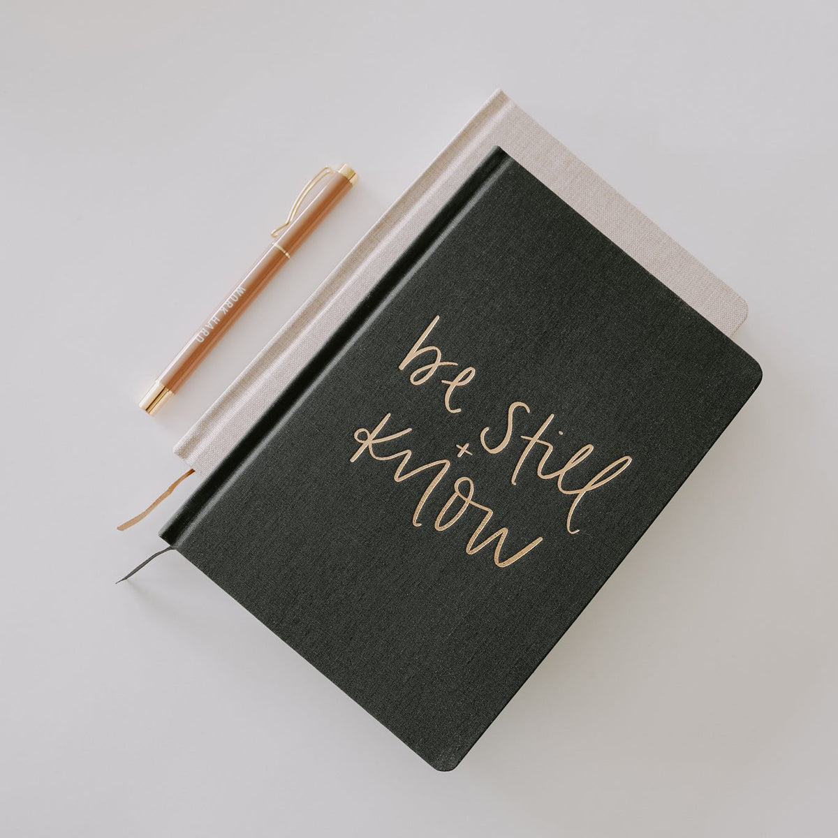 Black notebook with 'be still know' text and a pen on a light gray background