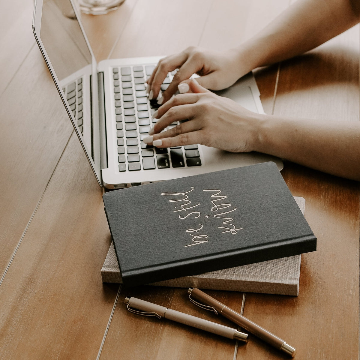 Person typing on a laptop with a notebook and pen on a wooden surface