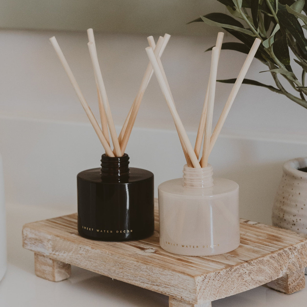 Two diffusers with wooden sticks on a wooden stand, one black and one beige, with a plant in the background.