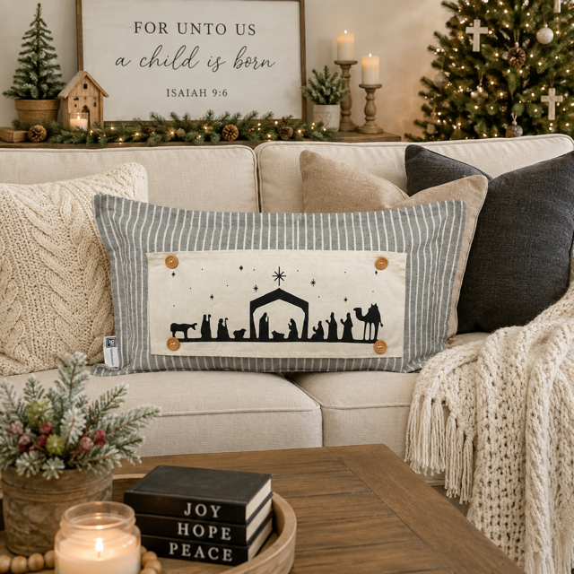 A rectangular Christmas throw pillow with a gray and white striped border and a central cream panel showing a black nativity scene, styled on a beige sofa in a decorated living room.