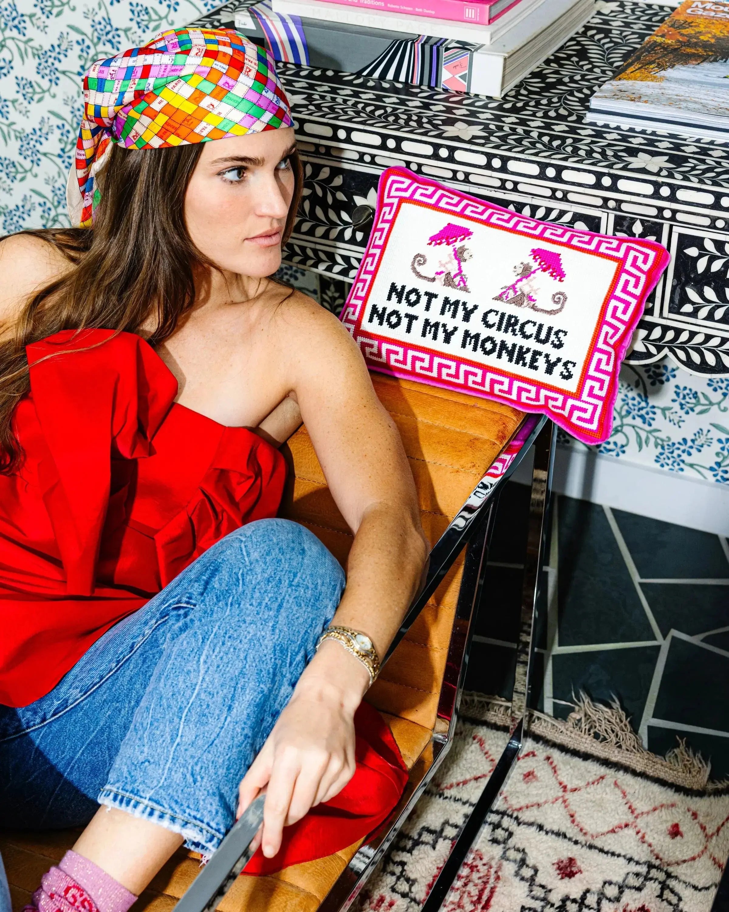 Woman in red top with colorful headscarf, beside 'Not My Circus Not My Monkeys' pillow on patterned chair