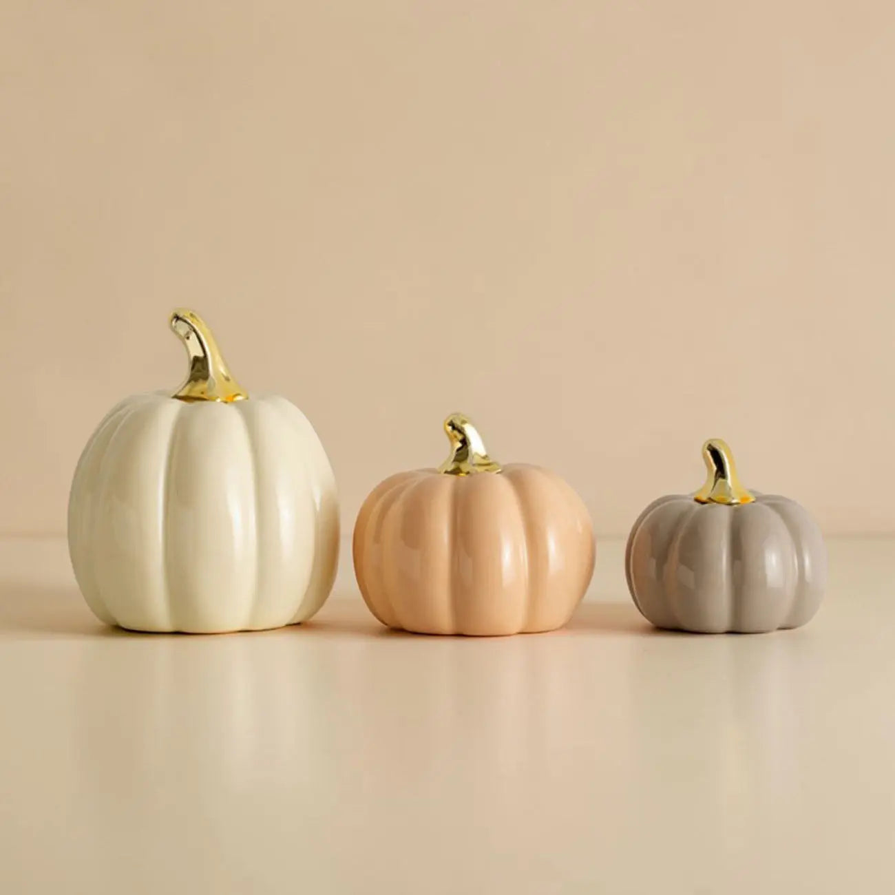 Three decorative pumpkins in white, pink, and gray with gold stems on a beige background