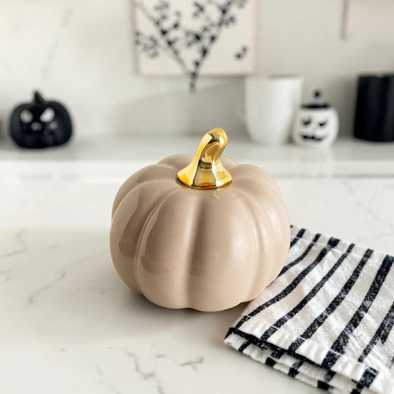 Beige ceramic pumpkin with gold stem on a marble surface, with Halloween decorations in the background.
