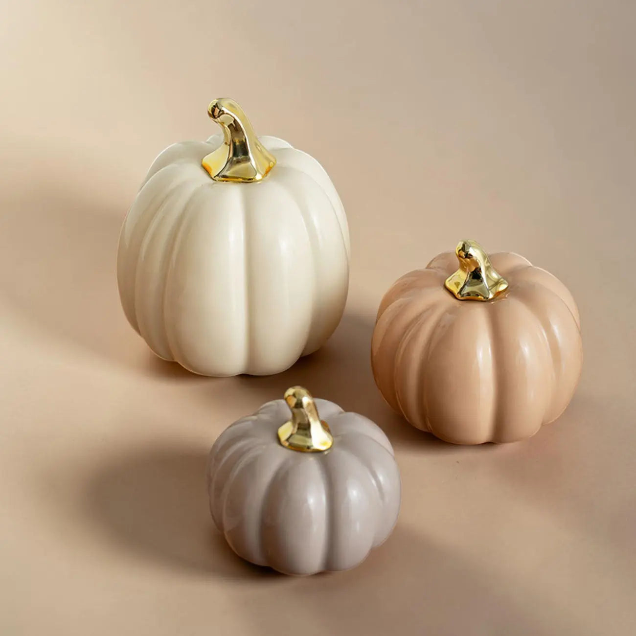 Three decorative pumpkins in white, beige, and gray with gold stems on a beige background.