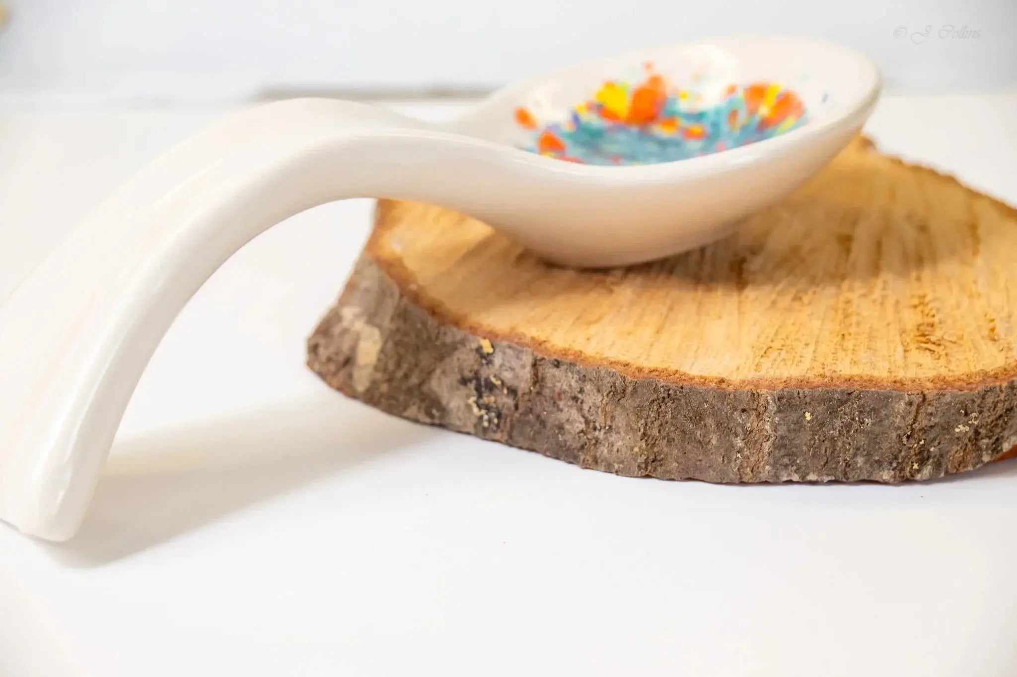 White ceramic spoon rest with colorful speckled design, displayed on rustic wood slice for farmhouse kitchen decor