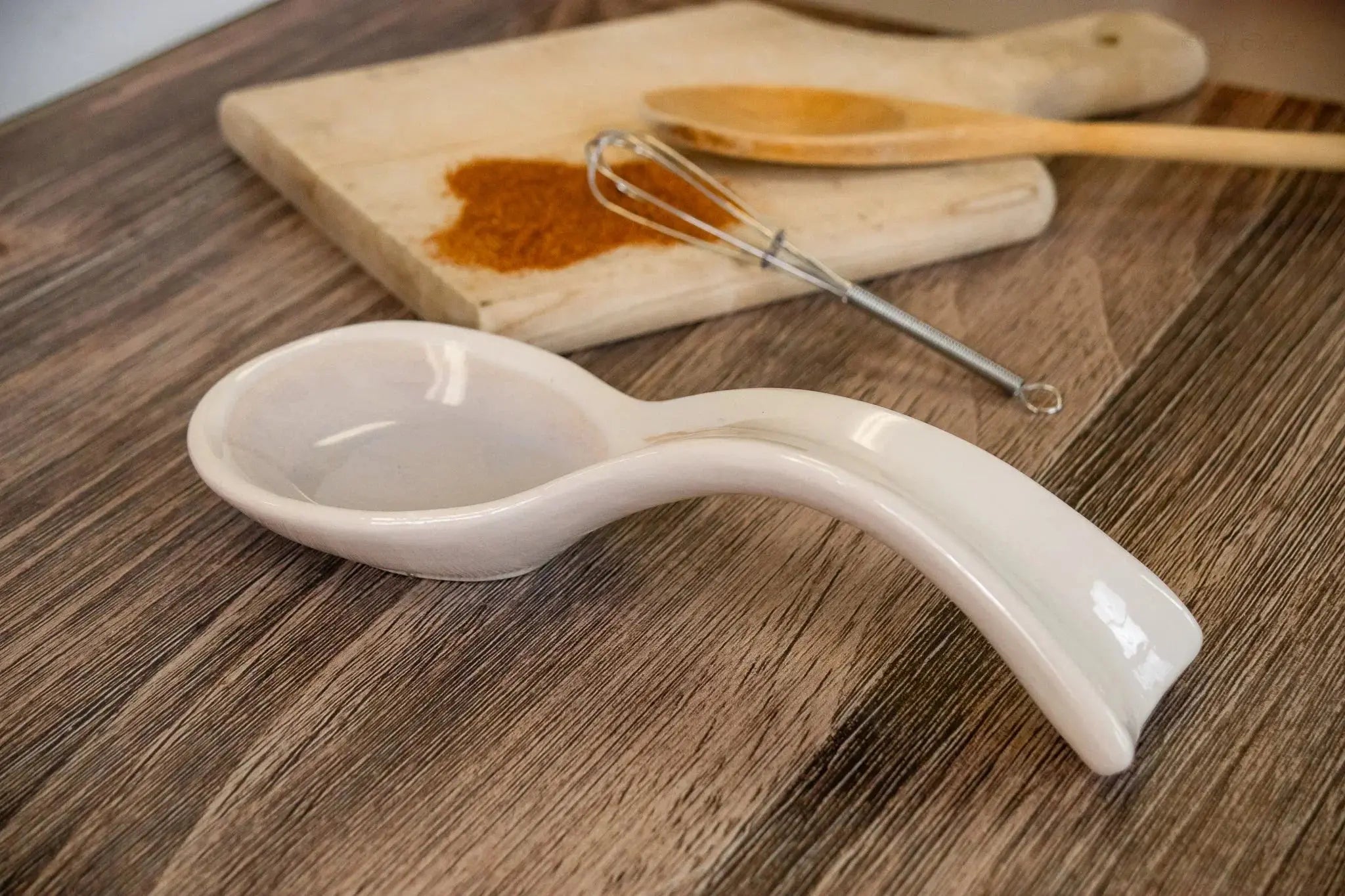 White ceramic spoon rest on wood table with cutting board, whisk, and spice