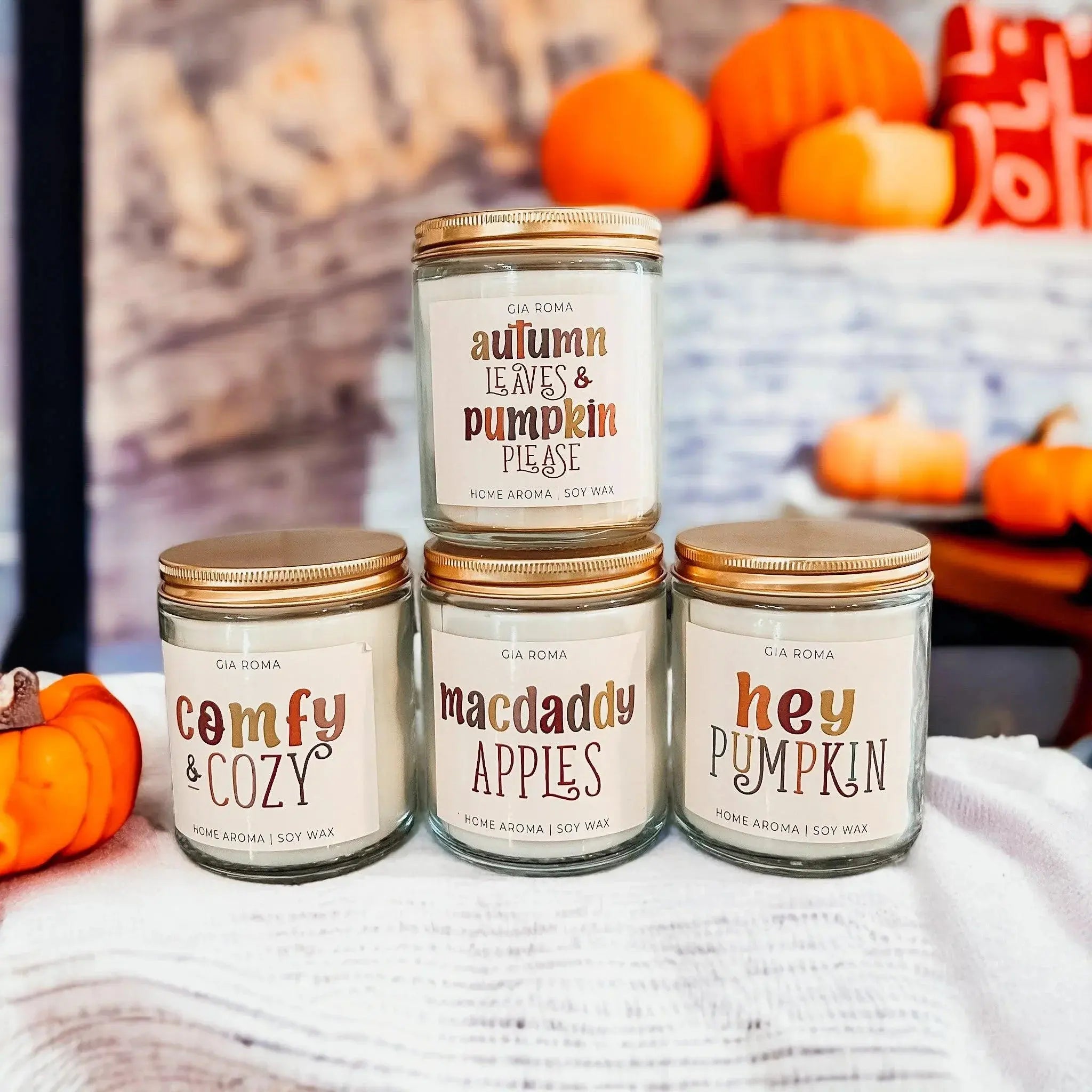 Five candles with autumn-themed labels on a white surface with pumpkins in the background