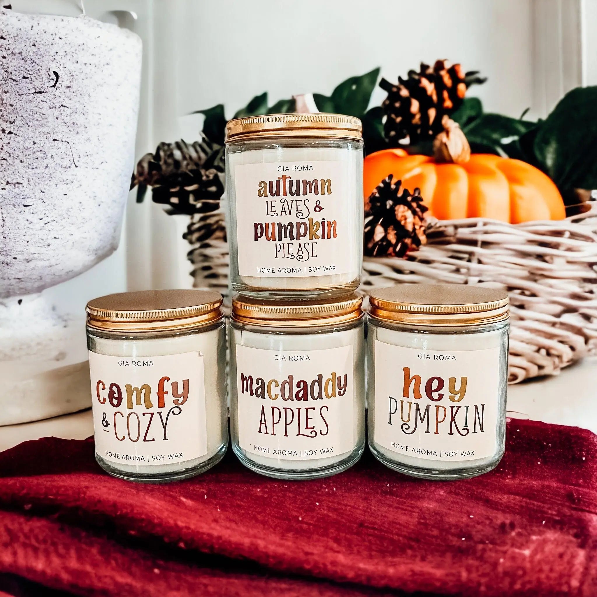 Four candles with autumn-themed labels on a red blanket with pumpkins and pinecones in the background.