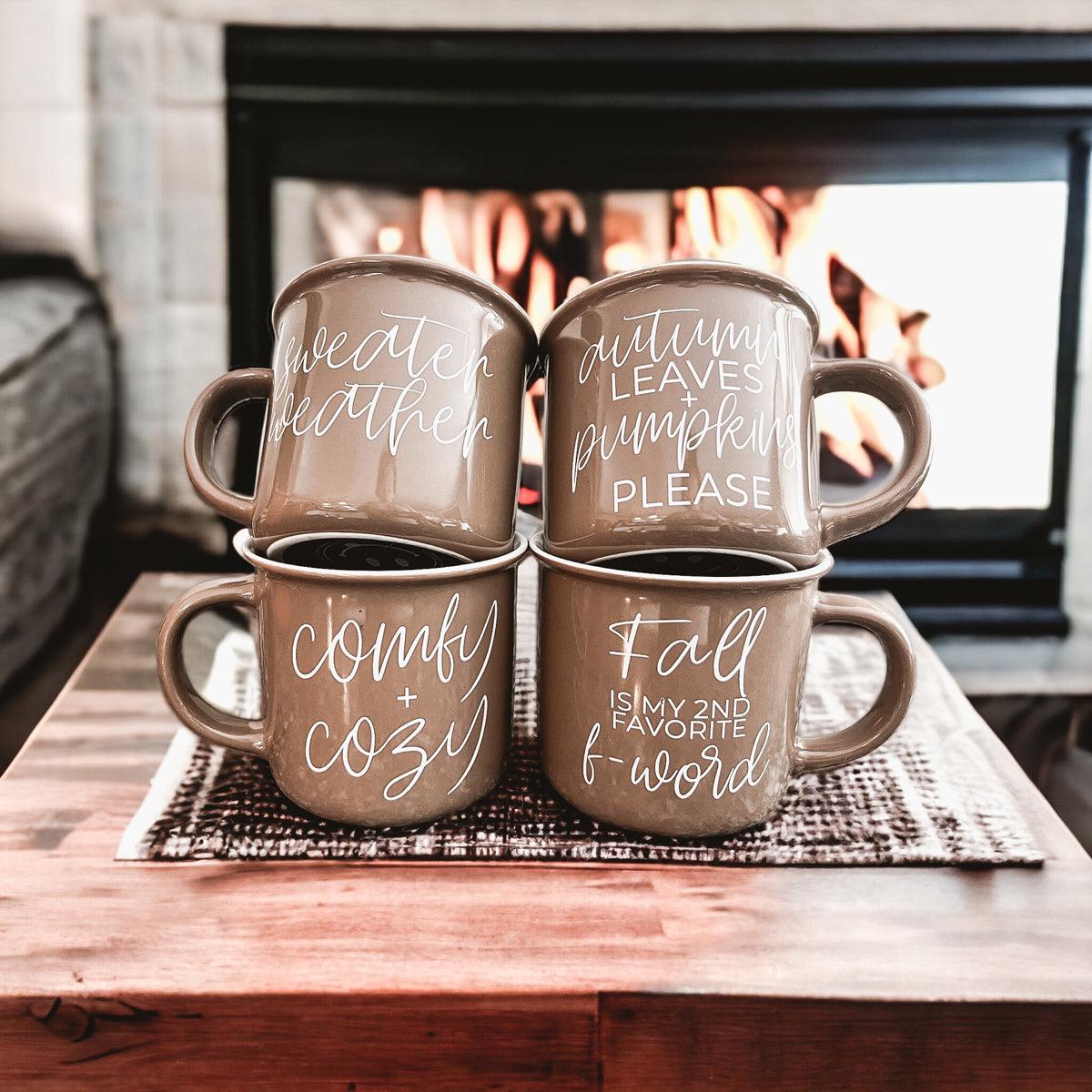 Four brown mugs with autumn-themed text on a wooden table in front of a fireplace.