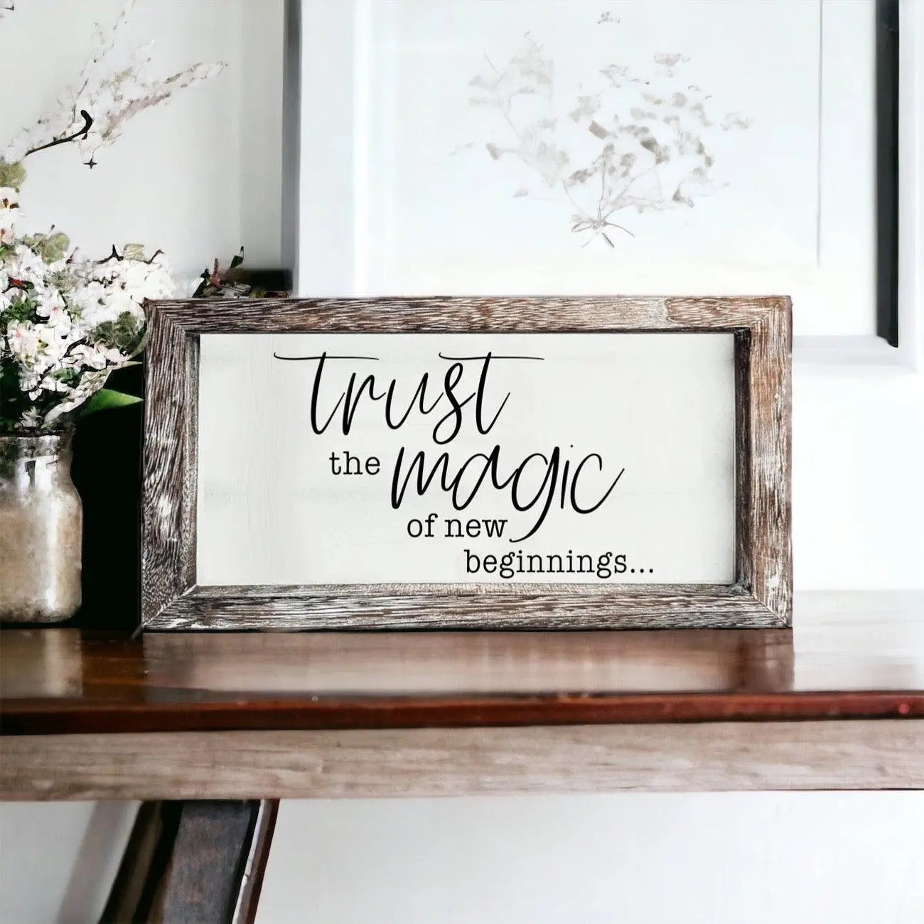 Framed sign with 'Trust the magic of new beginnings' text on a wooden surface with flowers in the background.