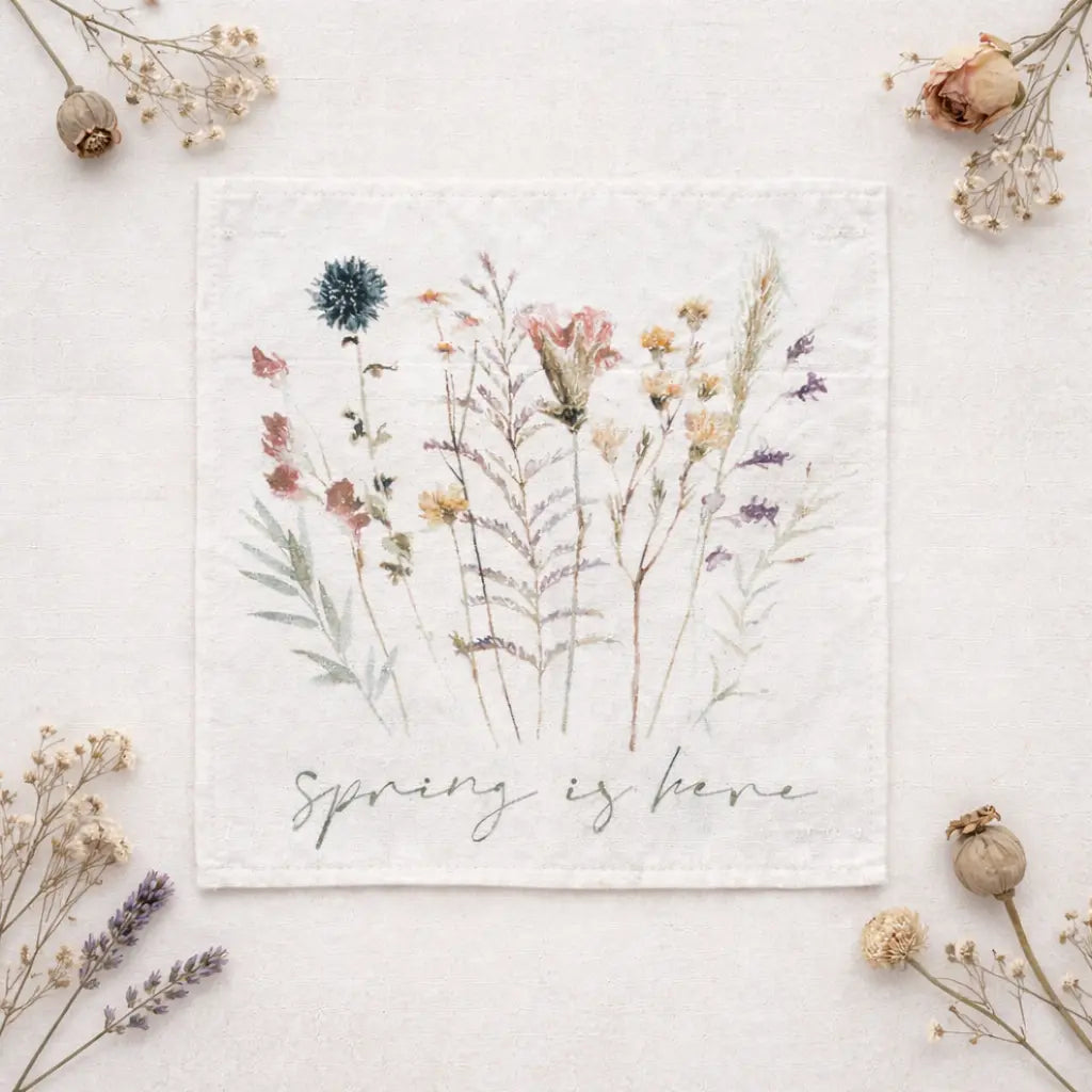 A square white fabric napkin or small cloth featuring a watercolor-style floral illustration with the words 'Spring is here' in script, styled flat-lay on a light neutral surface with dried flowers around it.