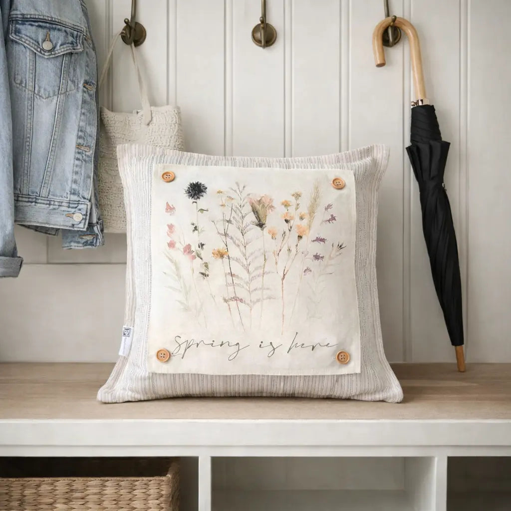 A square decorative throw pillow in a ribbed cream fabric with a central printed panel showing wildflowers and the words 'Spring is here,' styled on a wooden bench in a home interior with a jacket, bag, and umbrella nearby.