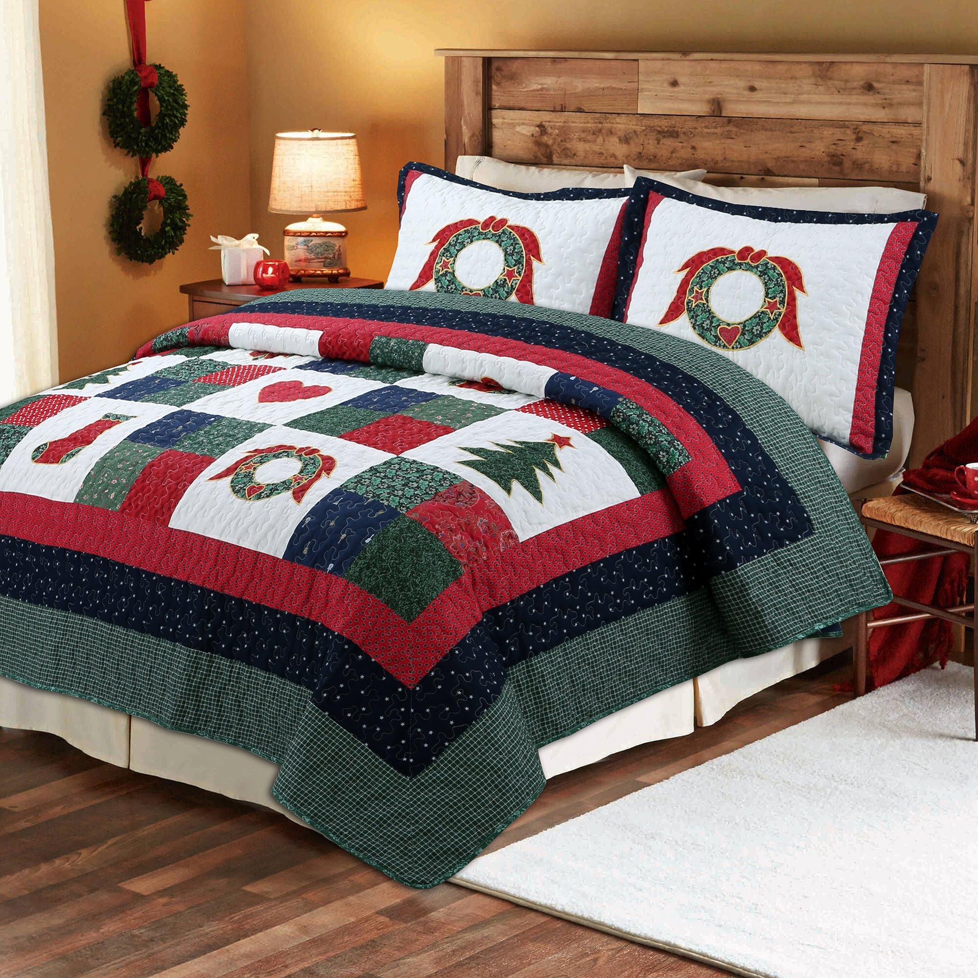 A Christmas-themed quilted bedding set on a bed, featuring a patchwork design with red, green, and white sections, a Christmas tree motif, stockings, and wreaths, styled in a warm bedroom setting.