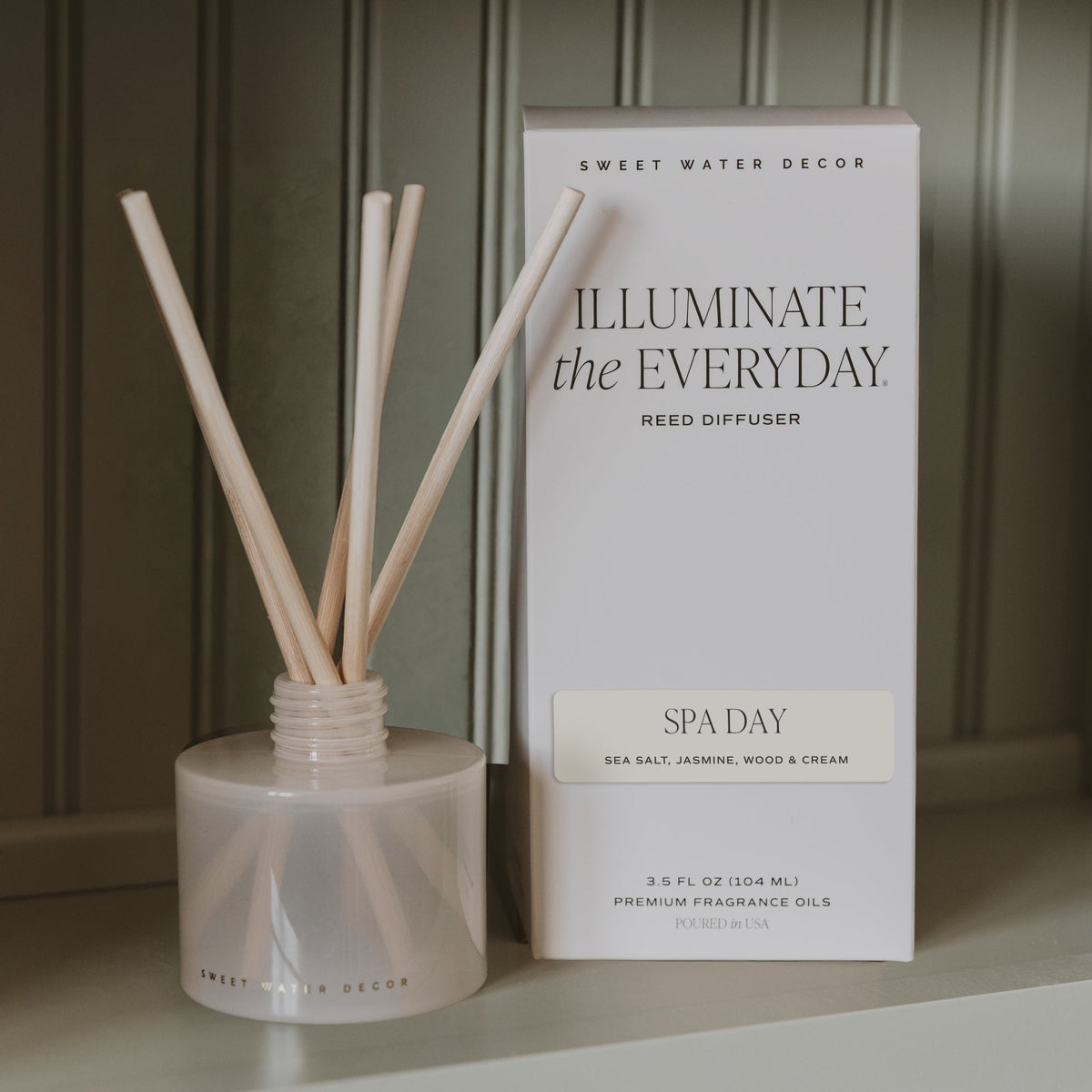 A reed diffuser bottle with pale natural reeds beside its white box. The box reads 'SWEET WATER DECOR', 'ILLUMINATE the EVERYDAY', and 'SPA DAY'.