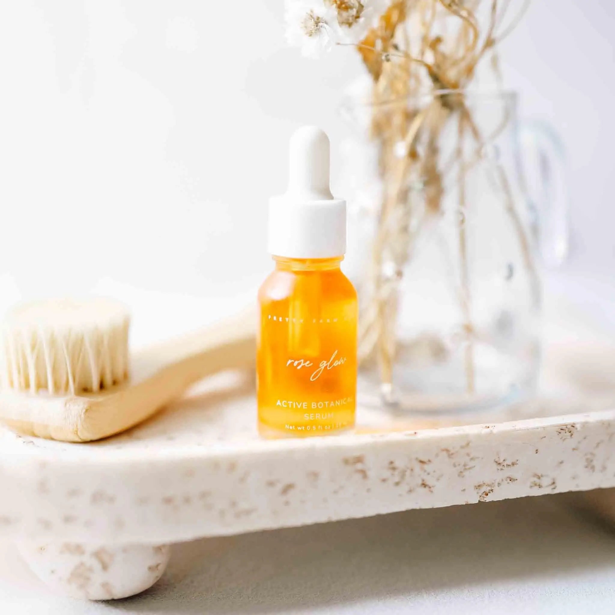Rose Glow active botanical serum in amber glass bottle with dropper, on rustic tray with brush and dried flowers