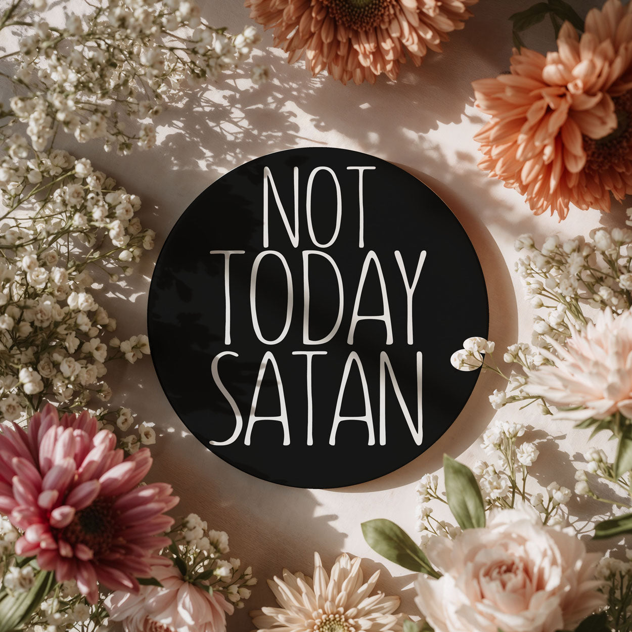 Circular sign with 'Not Today Satan' text surrounded by flowers on a light background