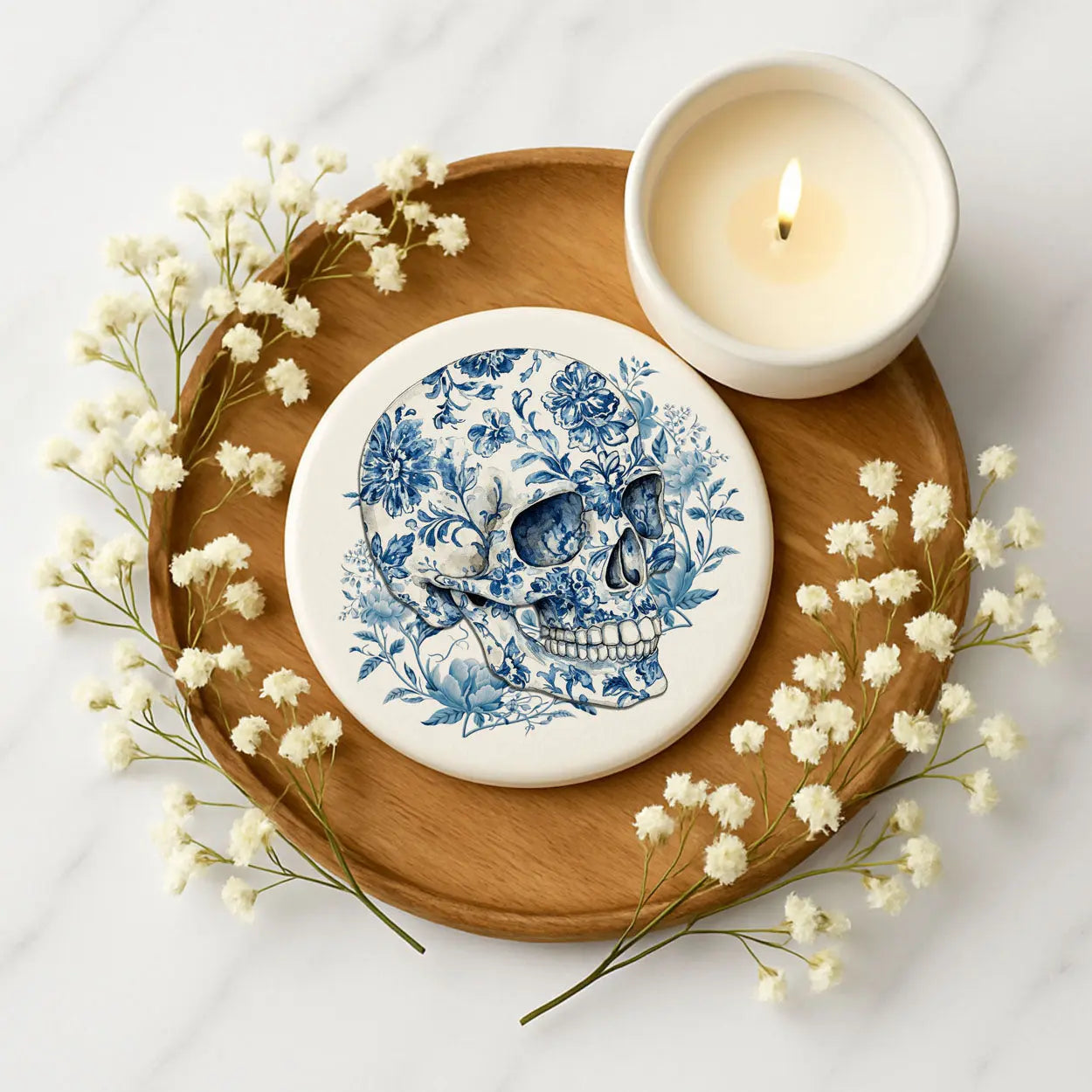 Decorative skull with floral patterns on a stone coaster, surrounded by white flowers and a lit candle on a wooden tray.