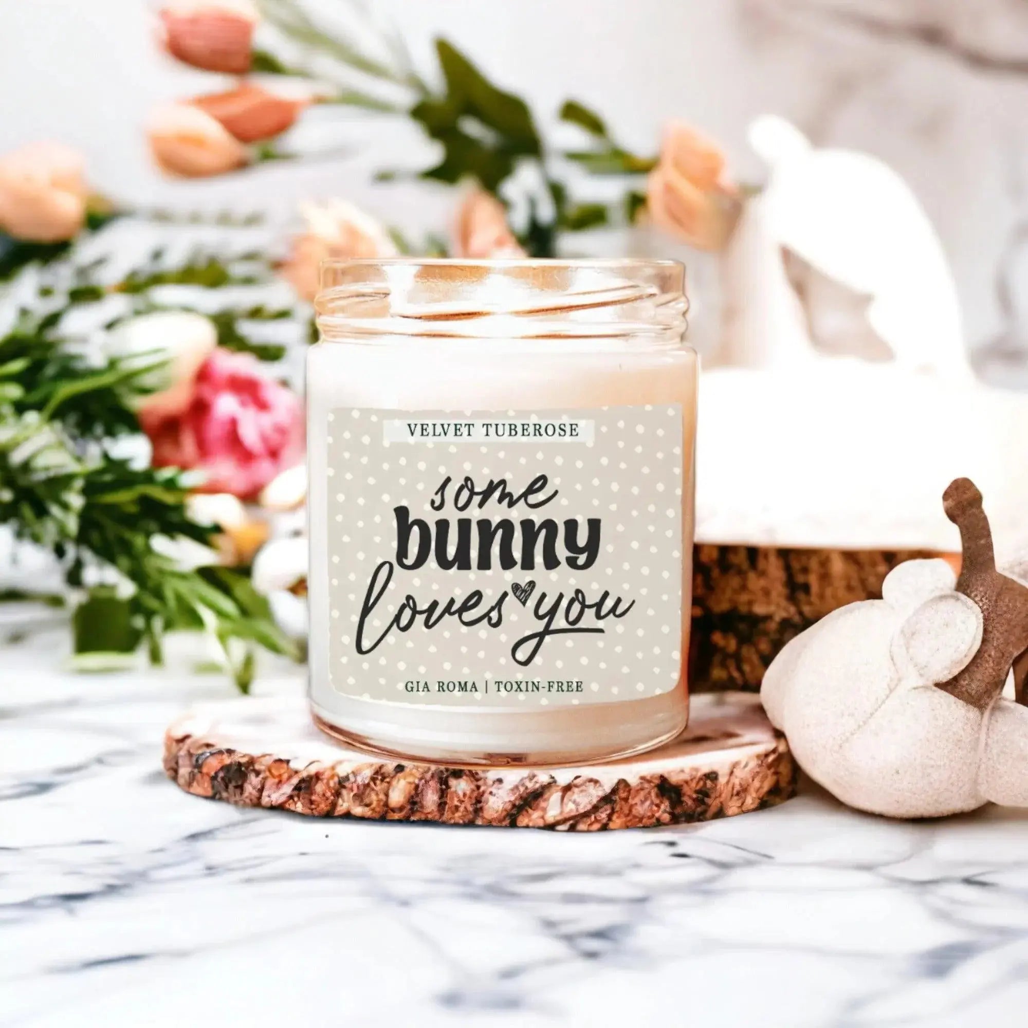 Candle with 'some bunny loves you' text on a marble surface with flowers and pumpkins in the background