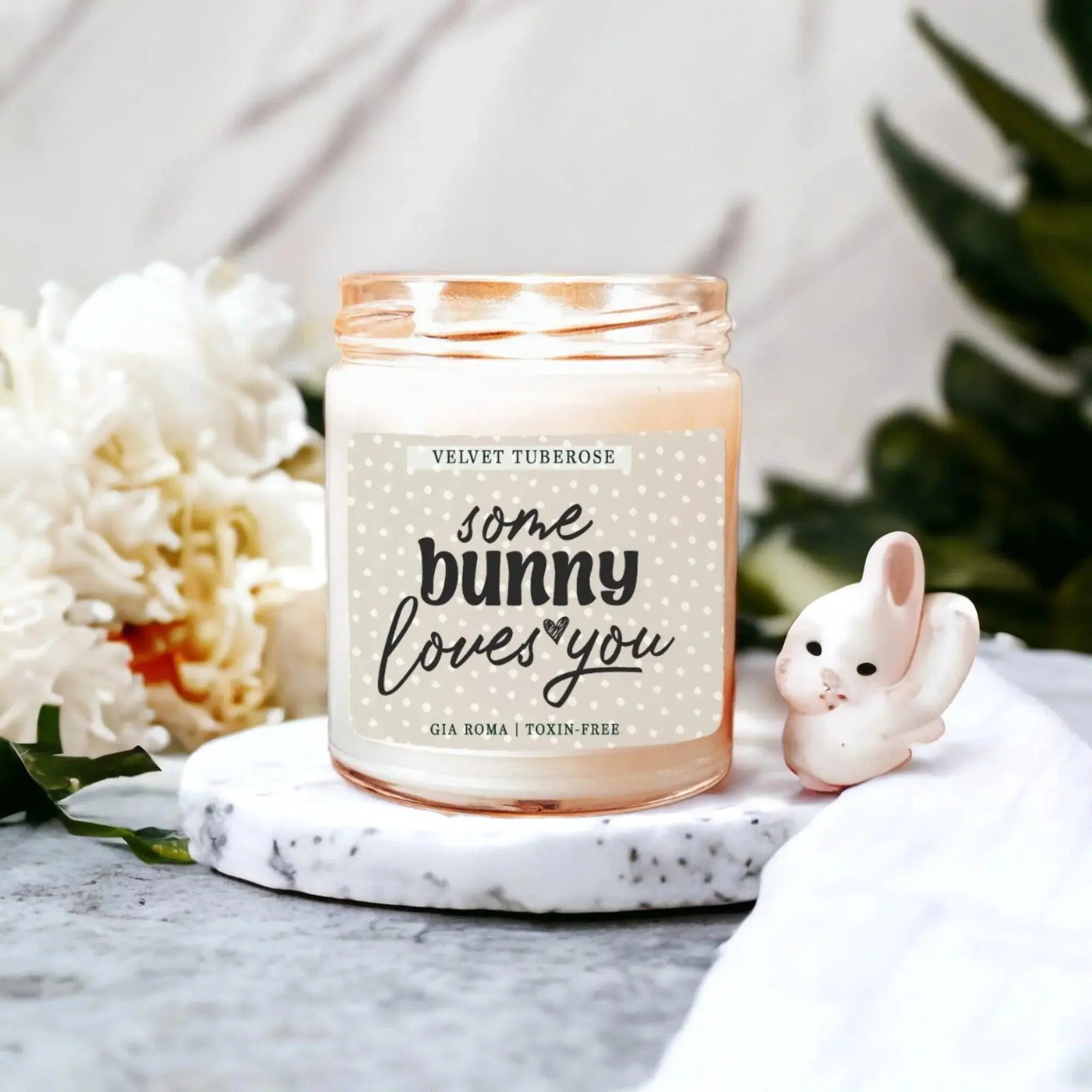 Candle jar with 'some bunny loves you' text, decorative rabbit, and flowers on a marble surface.