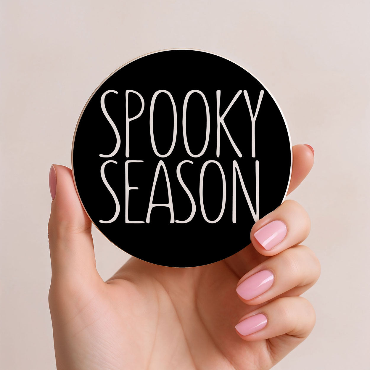 Hand holding a round black sticker with 'Spooky Season' text on a beige background