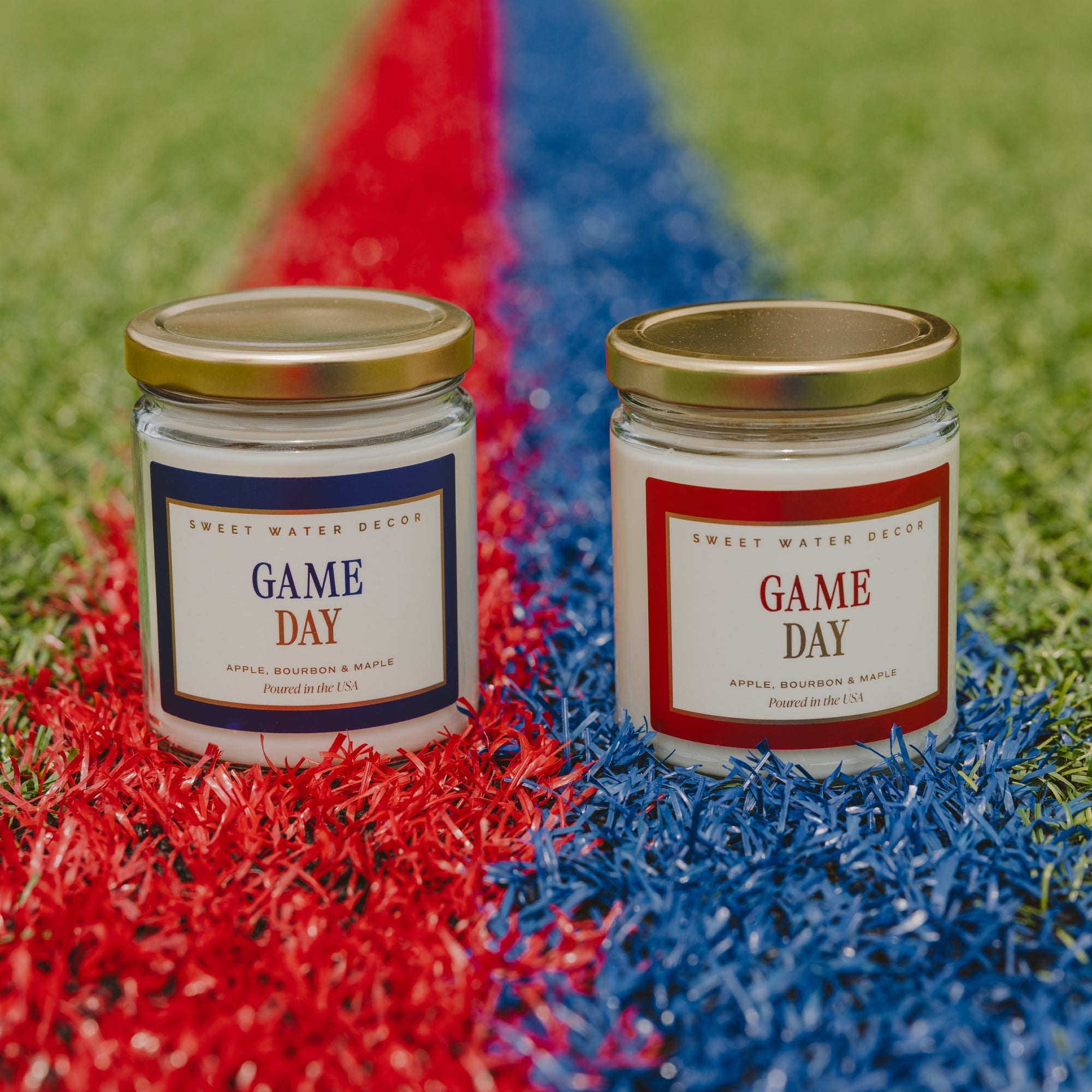 Two glass jar candles labeled 'GAME DAY' by Sweet Water Decor are placed on artificial turf over red and blue stripes, with green turf in the background.
