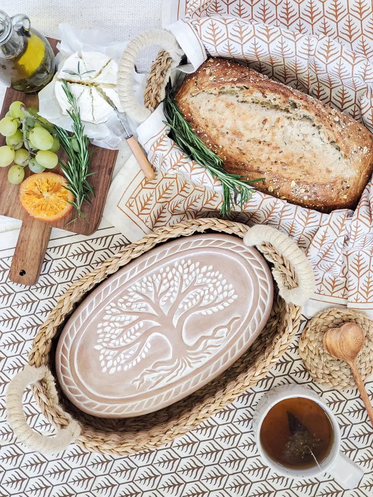 Cozy farmhouse bread basket with artisan loaf, cheese, grapes, and tree-of-life ceramic tray on leaf-patterned cloth