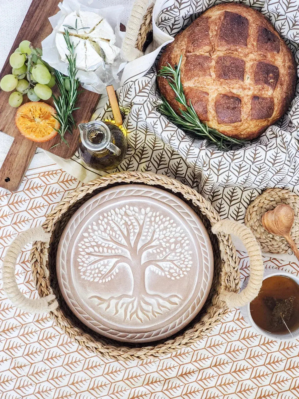 Rustic farmhouse tray with tree of life design, artisan bread, cheese, grapes, and rosemary on leaf-patterned cloth
