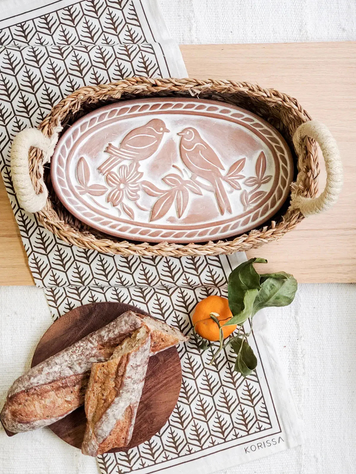 Woven tray with bird-patterned ceramic platter, rustic bread, and orange on leaf-print cloth for cozy farmhouse decor