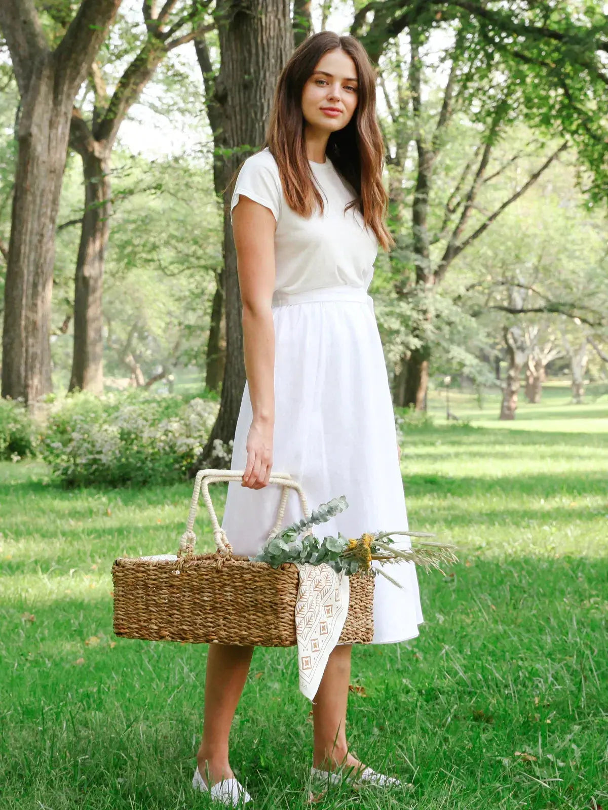 Woman in white dress holding woven picnic basket with eucalyptus, styled for cozy farmhouse decor