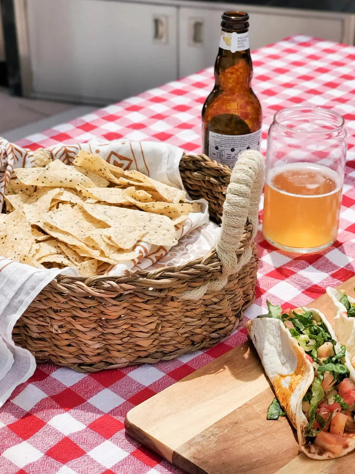 Woven basket of tortilla chips, tacos on wood board, beer bottle and glass on red checkered tablecloth