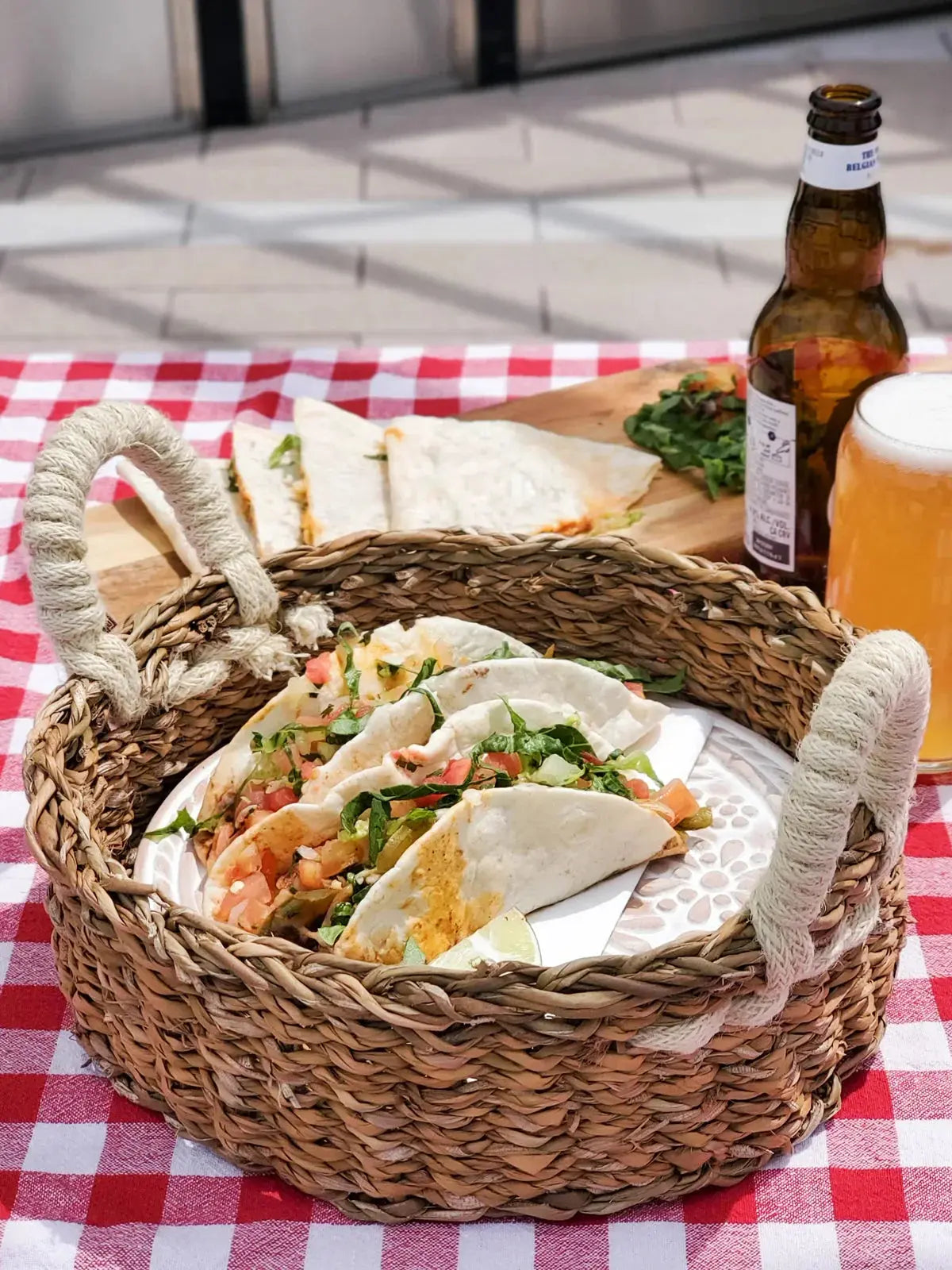 Woven basket with fresh tacos on red checkered picnic table, beer bottle and glass nearby