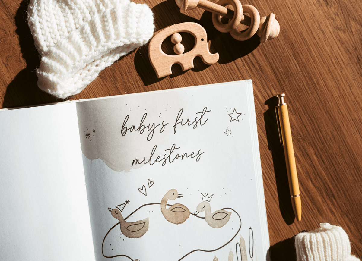 Notebook titled 'baby's first milestones' with baby items on a wooden surface