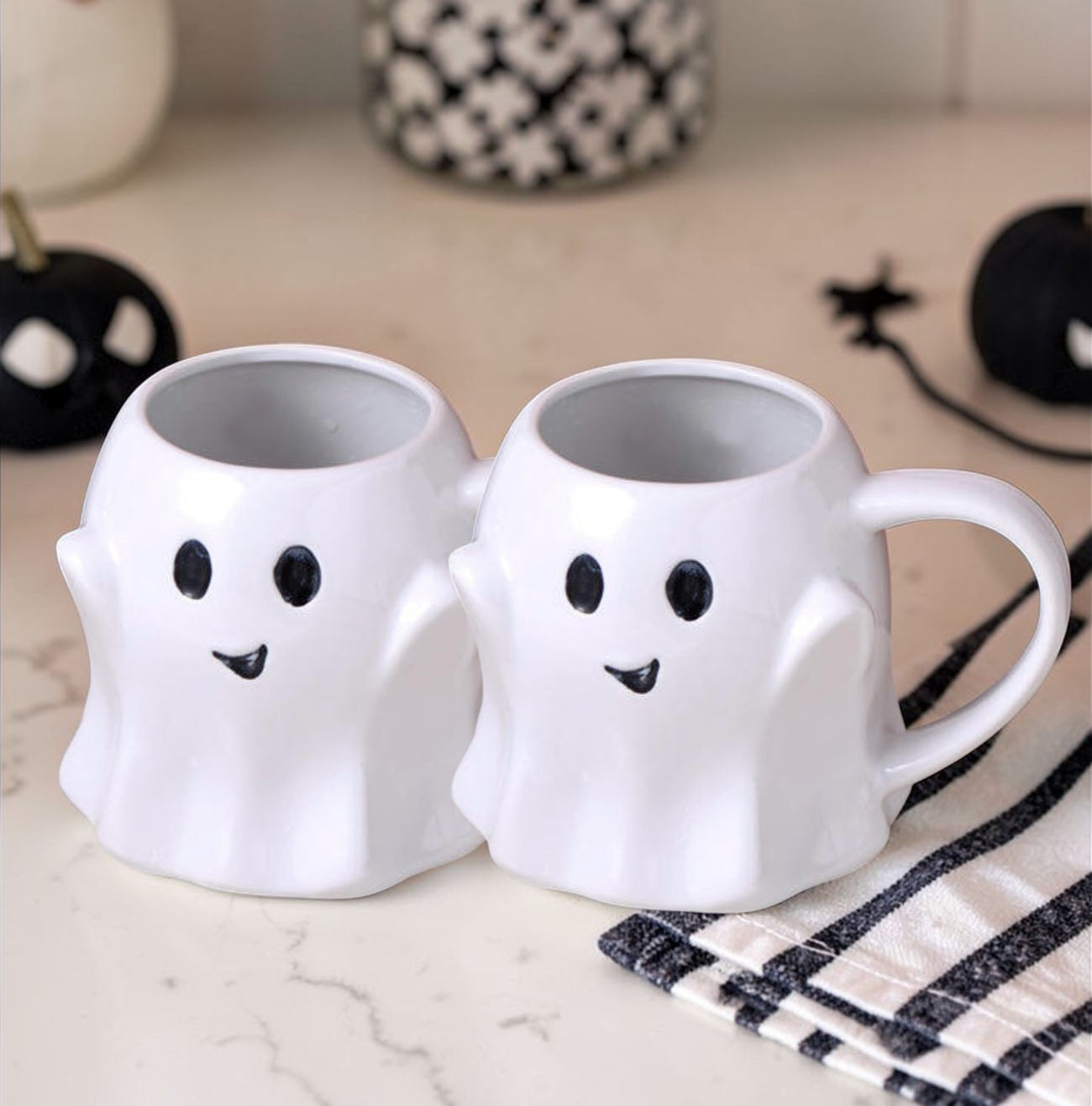 Two ghost-shaped mugs on a surface with Halloween decorations.