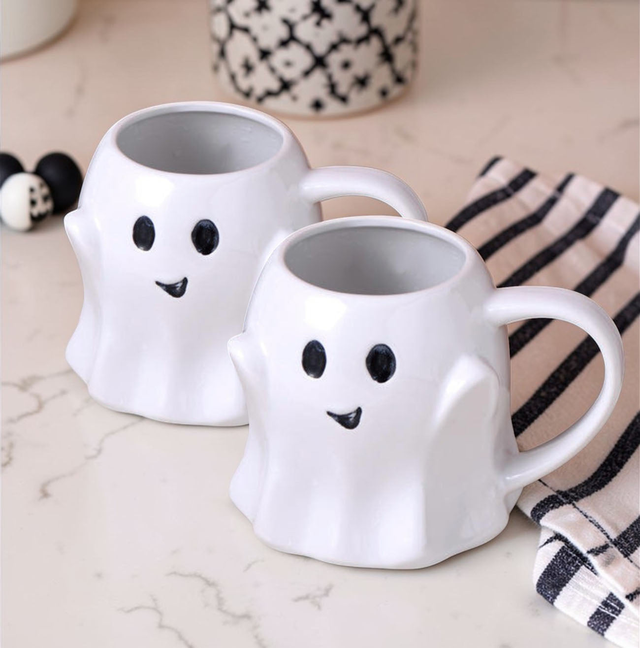 Two ghost-shaped mugs on a marble surface with decorative items in the background.