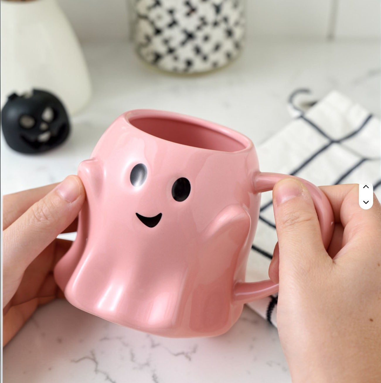 Pink ghost-shaped mug held by hands on a marble surface with a blurred background