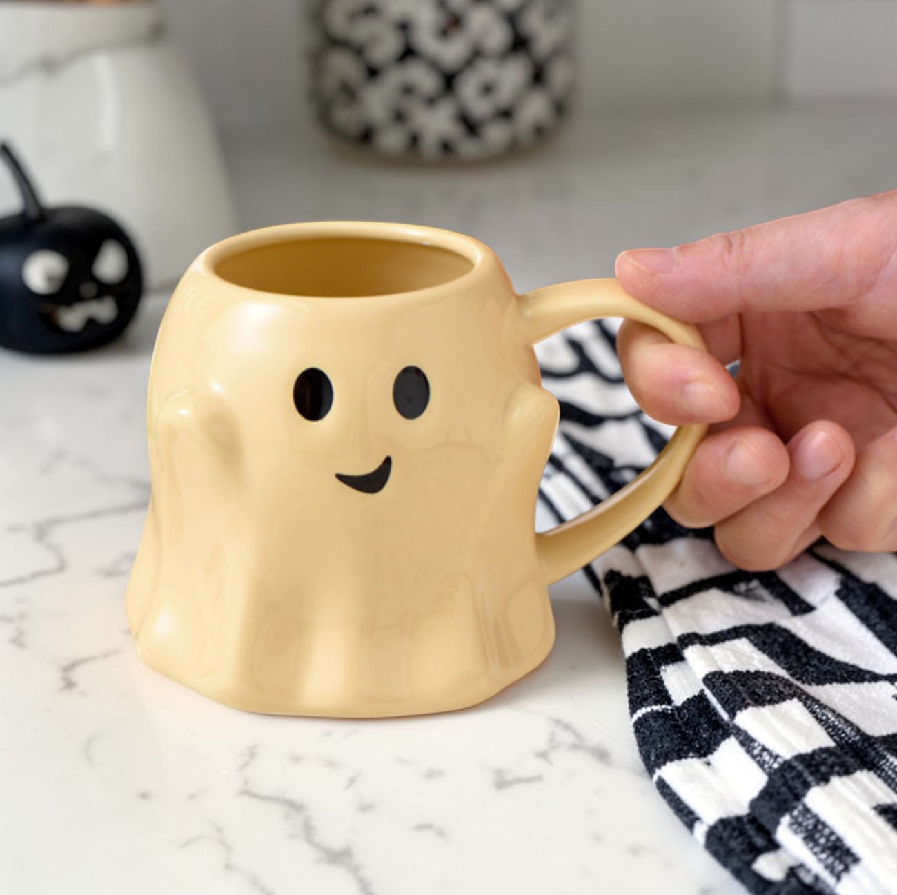 Hand holding a ghost-shaped mug on a marble surface with Halloween decorations in the background.