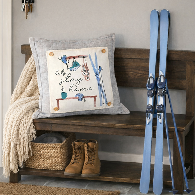 Wooden bench with a decorative pillow, boots, and skis against a neutral wall.