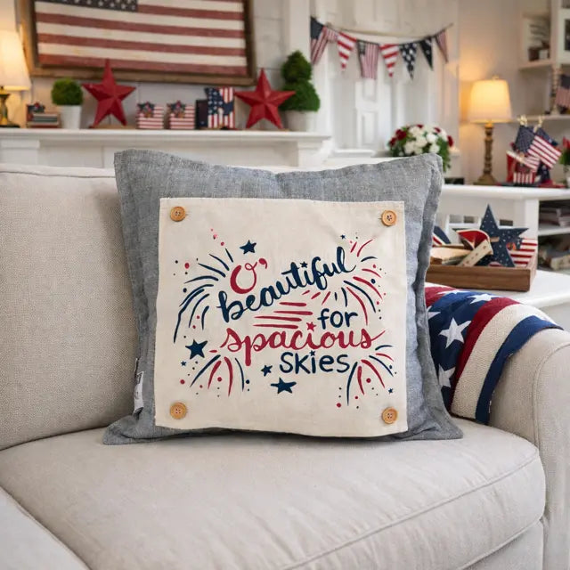 A square throw pillow with a gray linen-textured border and a cream center panel printed with the phrase 'Beautiful for Spacious Skies' in red and navy lettering with fireworks and stars. The pillow is styled on a light beige sofa in a decorated living room with patriotic themes.