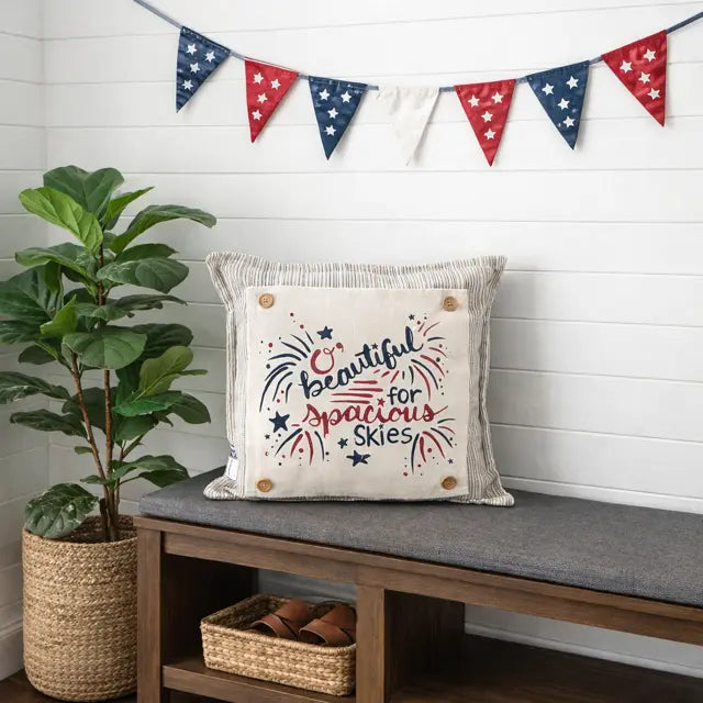 Decorative pillow with 'Beautiful for Spacious Skies' text on a bench with a plant and flag banner in the background.