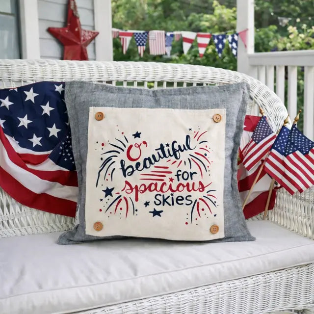 Decorative pillow with 'O' Beautiful for Spacious Skies' text on a white wicker chair with American flags.