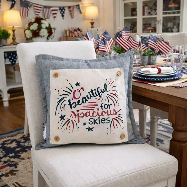 A square decorative throw pillow displayed on a white upholstered chair in a styled dining room. The pillow has a light blue-gray fabric border and a cream center panel with the phrase “Beautiful for Spacious Skies” in red and navy lettering, surrounded by red, white, and blue fireworks and star motifs, plus four wooden buttons in the corners.