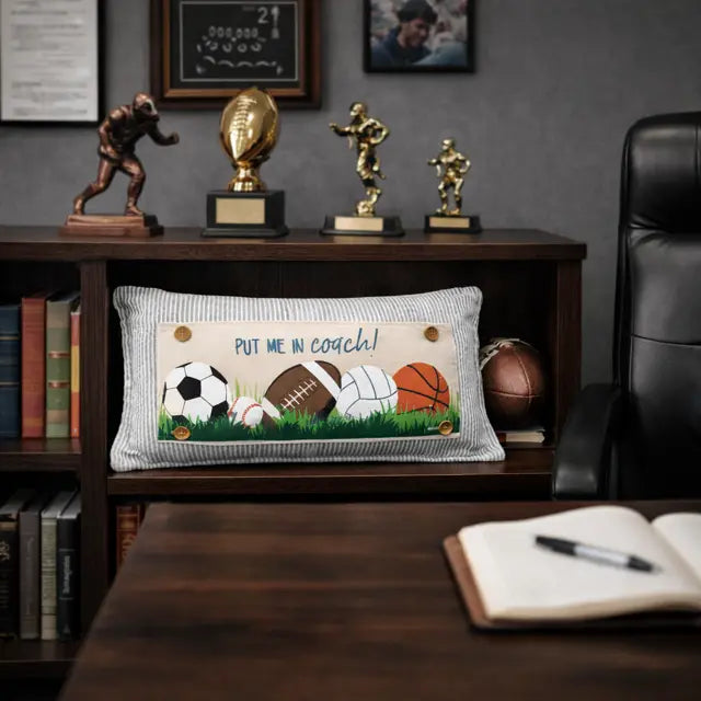 Decorative pillow with sports-themed design on a desk with trophies and books in the background.