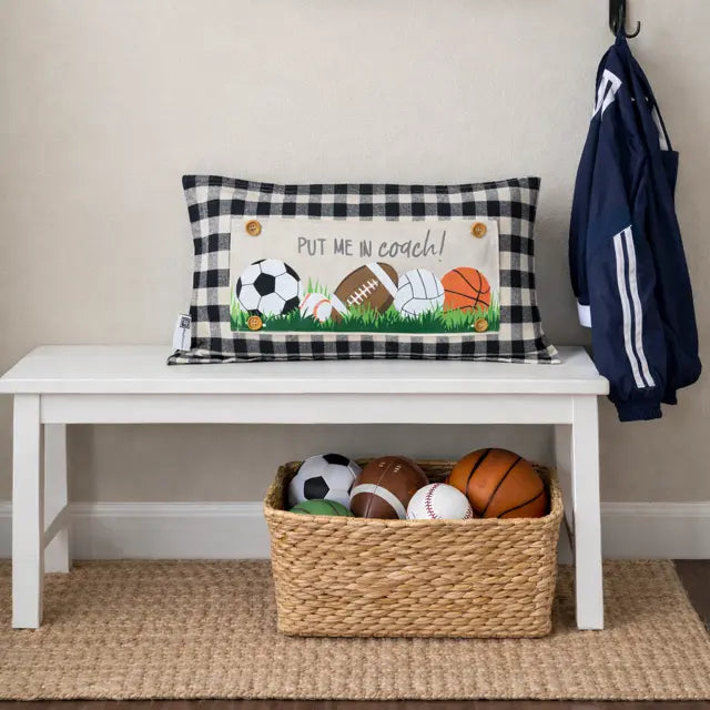 Decorative pillow with sports-themed design on a white bench with a basket of sports balls below.
